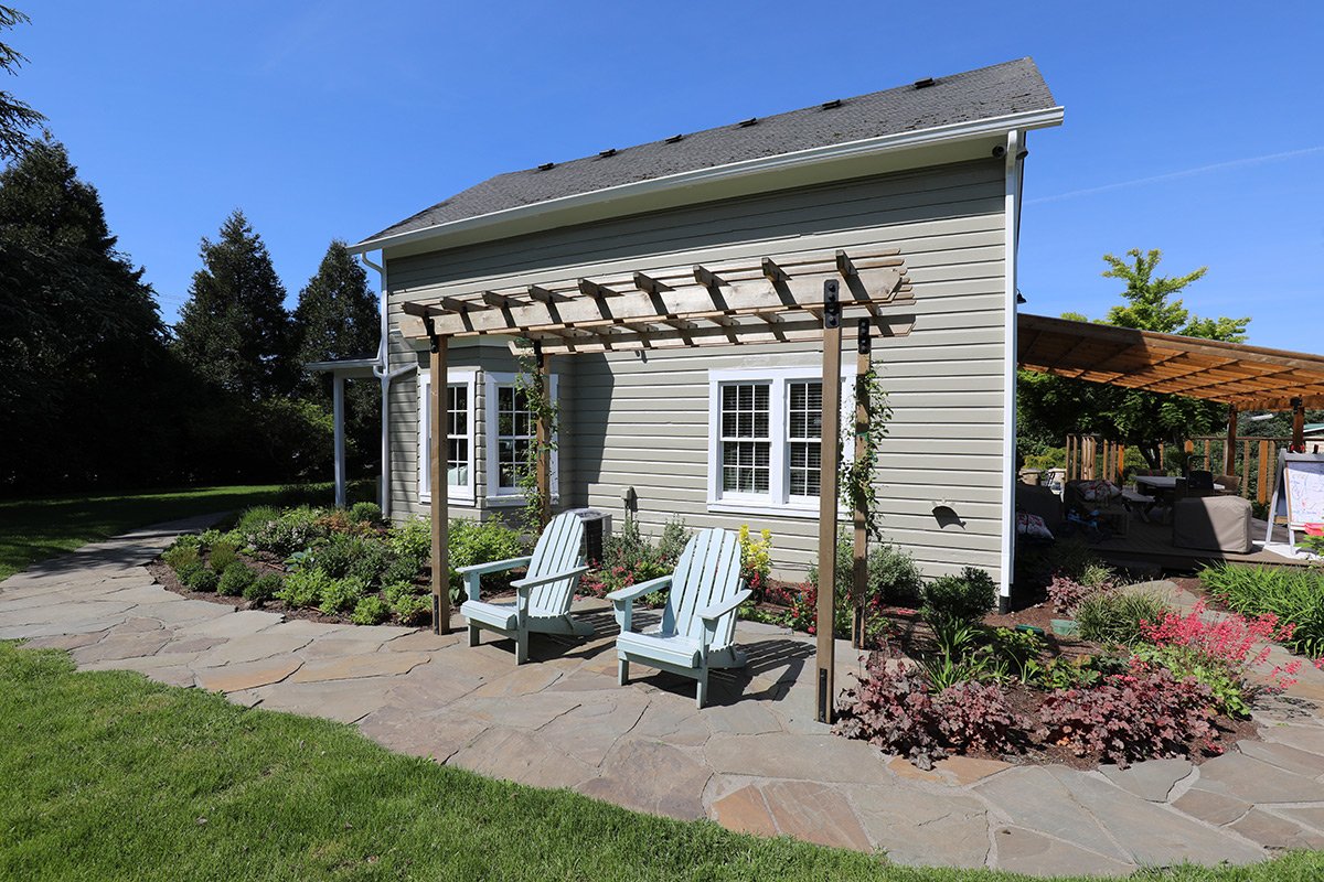 Backyard with a house, stone pathway, two Adirondack chairs under a wooden pergola, and a landscaped garden with flowers and greenery.