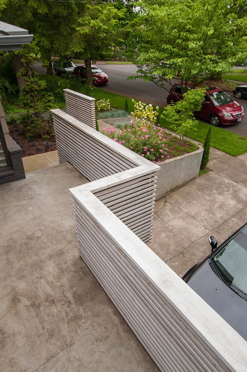View from a balcony showing a concrete patio, modern metal partition walls, a garden with pink and yellow flowers, green trees, parked cars on the street, and grass.
