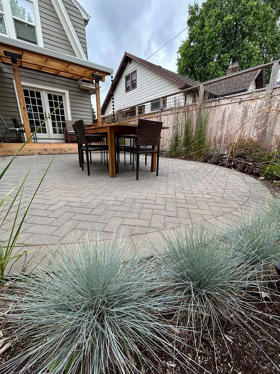 Backyard patio with a paved surface, outdoor dining table, and six chairs. There is a house with French doors and a small deck. A wooden fence borders the yard, with tall ornamental grass and trees nearby. Overcast sky.