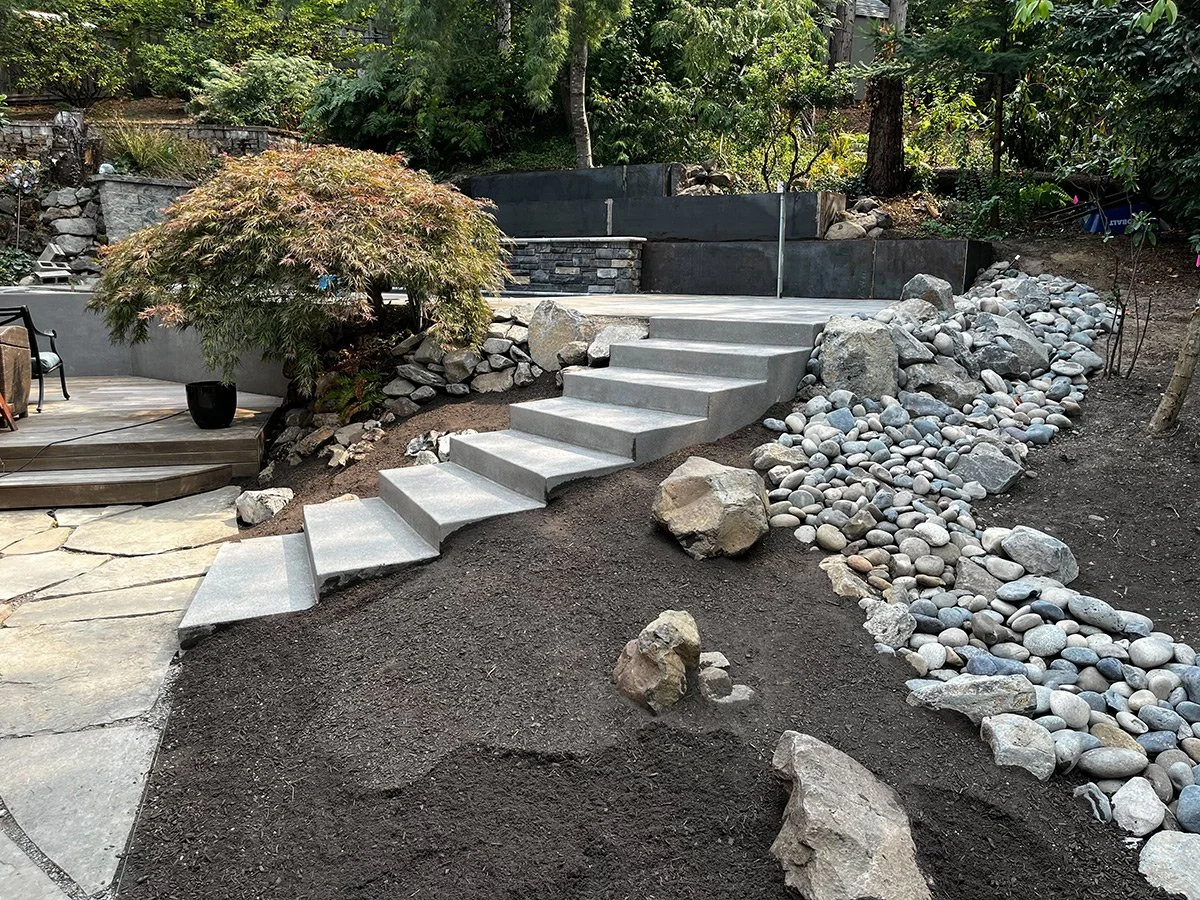 Backyard with concrete steps leading to a higher level, surrounded by rocks and a small gravel creek, with trees and shrubbery in the background.