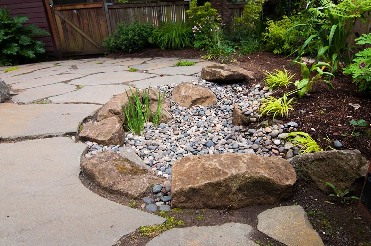 A garden pathway with irregular stone slabs surrounded by a landscaped garden bed with small rocks, bushes, and plants.