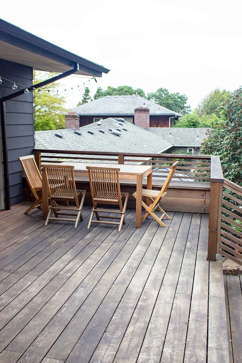 Wooden deck with a table and four chairs, overlooking neighboring rooftops and trees, during daytime.