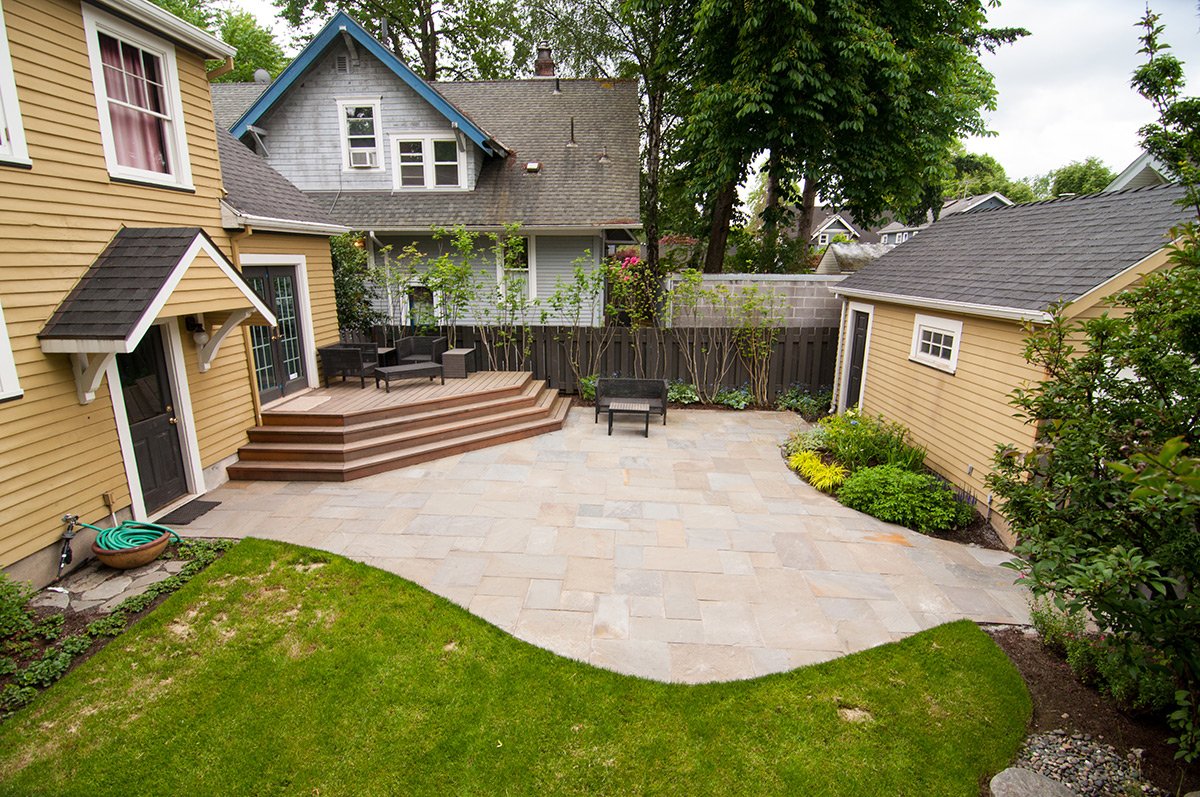 Backyard with a stone patio, small grassy area, wooden deck with outdoor furniture, and surrounding trees and shrubs.