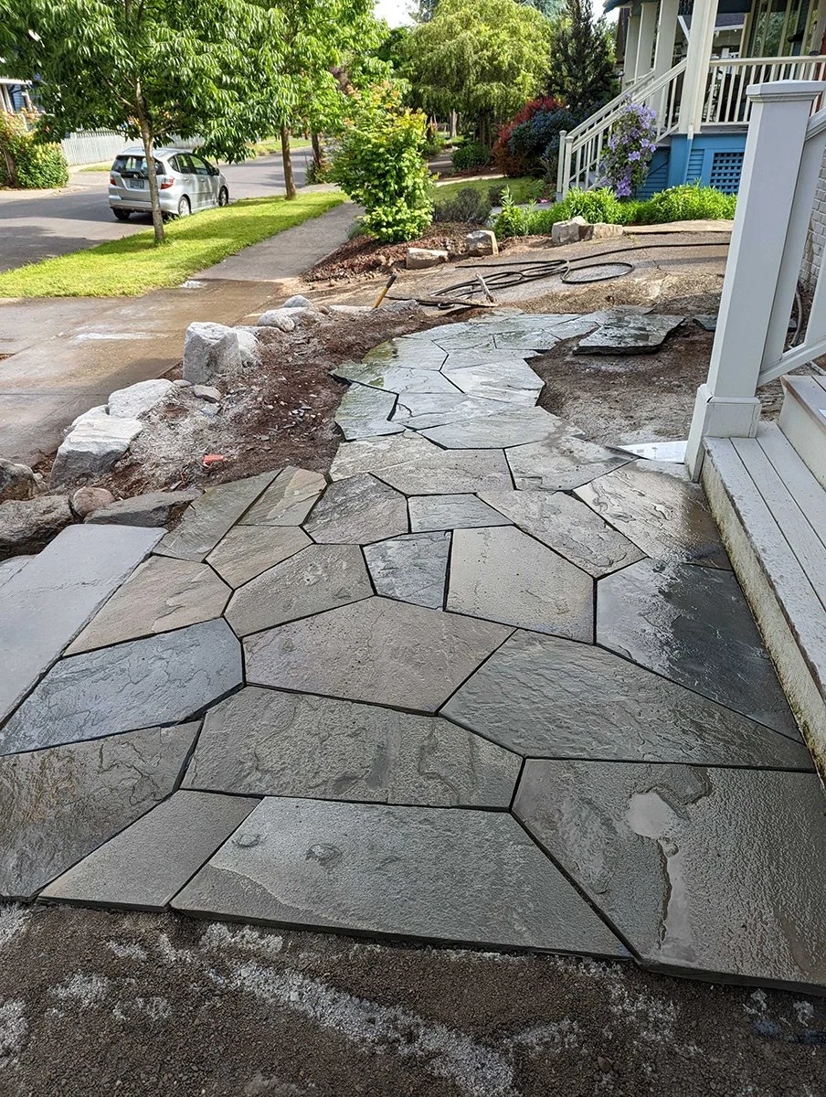 Front porch with newly installed irregular-shaped stone pathway leading to the sidewalk, with construction materials and tools nearby, and a residential street in the background with trees and parked cars.