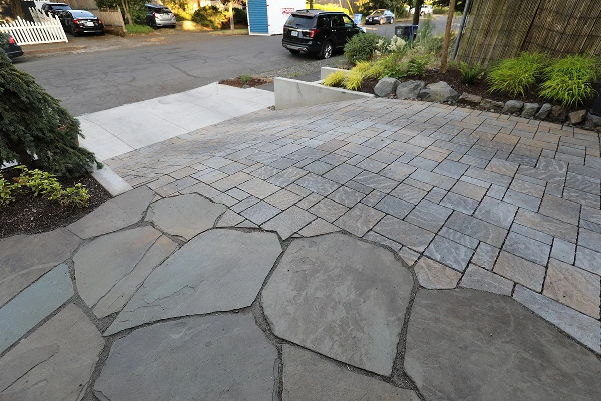 A paved outdoor area featuring stone and brick paving, a landscaped garden bed with plants and rocks, and a nearby parking lot with several cars.