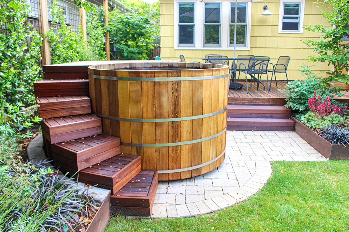 Backyard with wooden steps leading to a deck, a round hot tub, and patio furniture next to a yellow house with white-trimmed windows.
