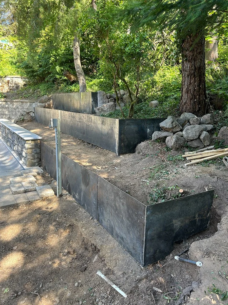 A garden with black metal wall planters, trees, rocks, and greenery.