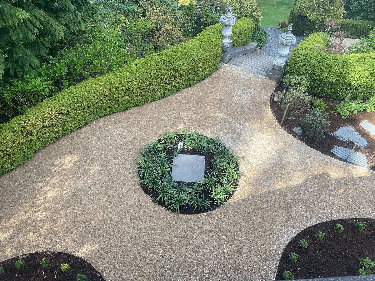 A landscaped garden with a gravel pathway, green shrubs, and ornamental planters with stairs and urns in the background.