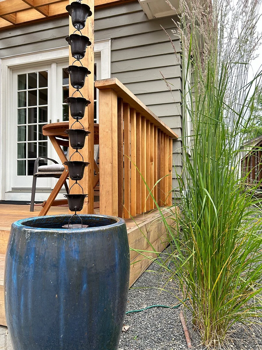 View of a backyard wooden porch with a railing, a chair, a sliding glass door, a tall plant, a blue ceramic water fountain with a tiered black metal fountain, and a house with gray siding.