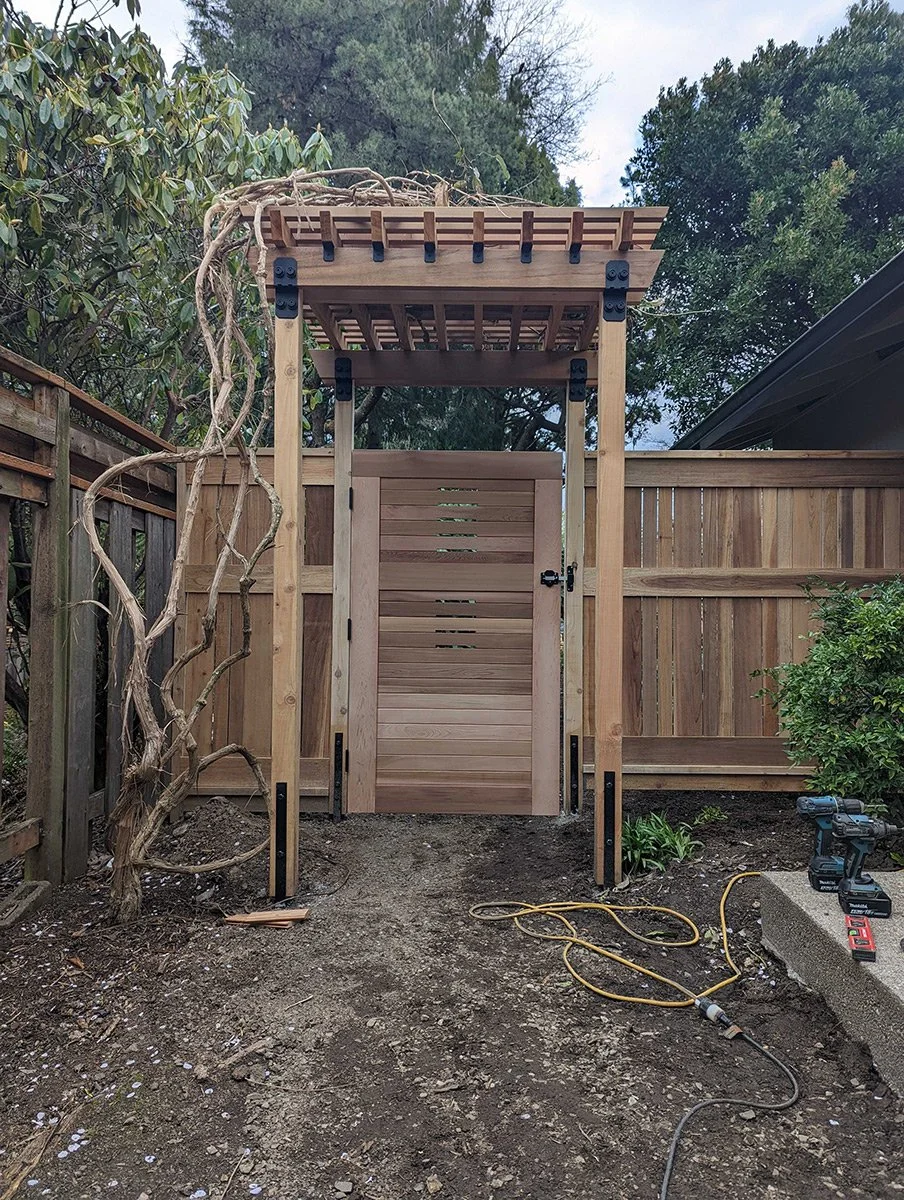 Wooden garden gate with an arched trellis top, installed within a backyard fenced area, with some power tools and an extension cord on the ground in front.