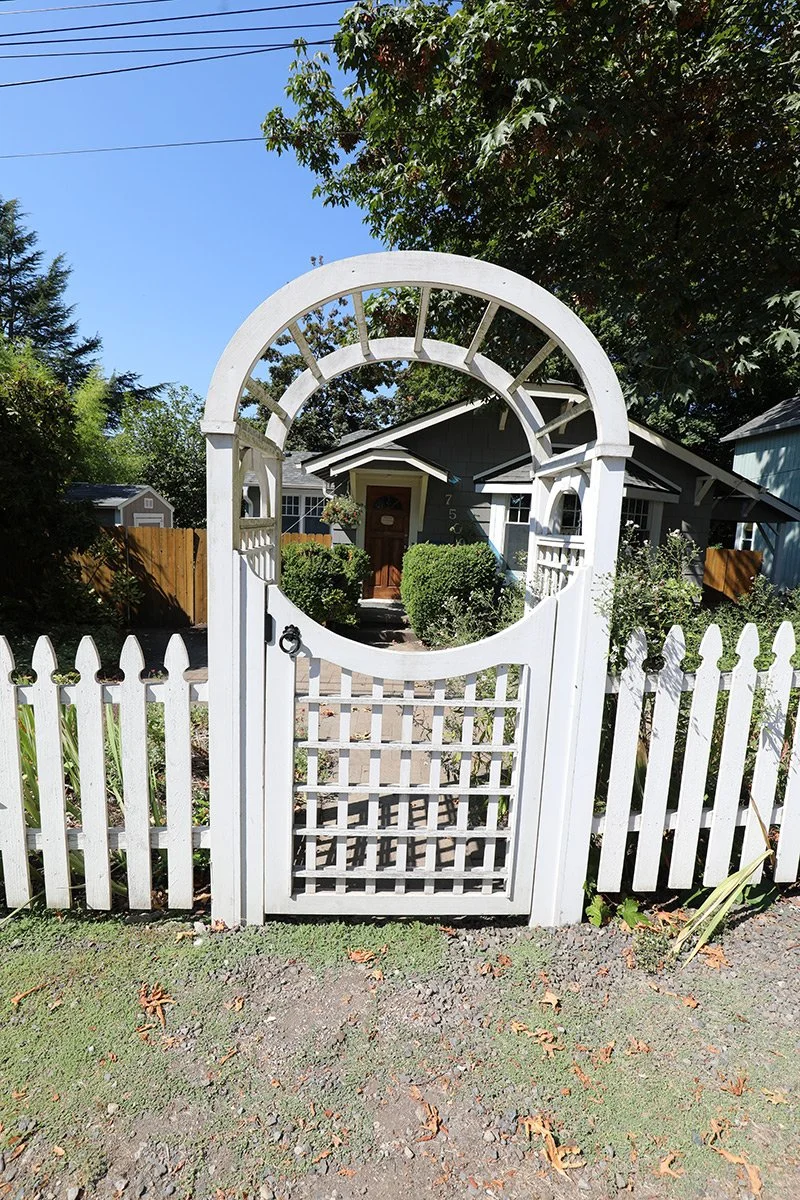 White garden gate with circular arch design leading to a front yard and house with a dark roof, surrounded by green bushes and trees.