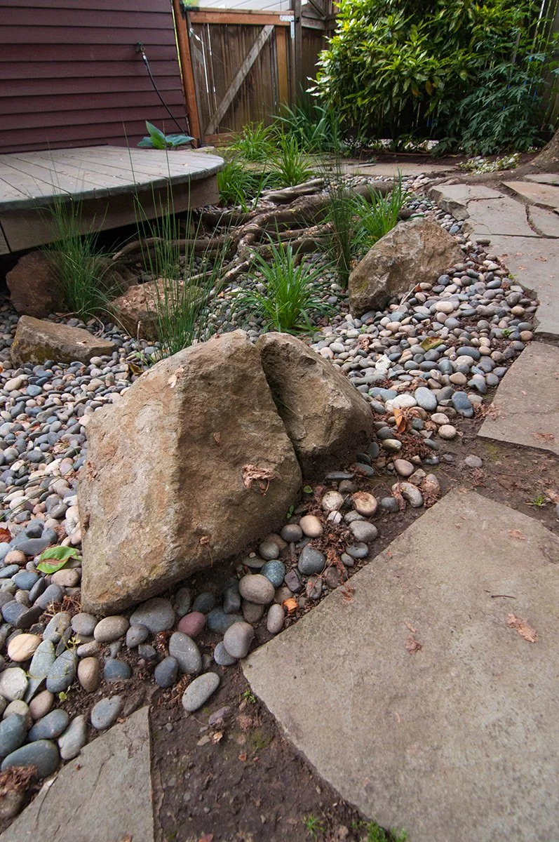 A landscaped backyard garden with large rocks, small pebbles, green plants, and a stone pathway next to a wooden deck and a wooden fence.