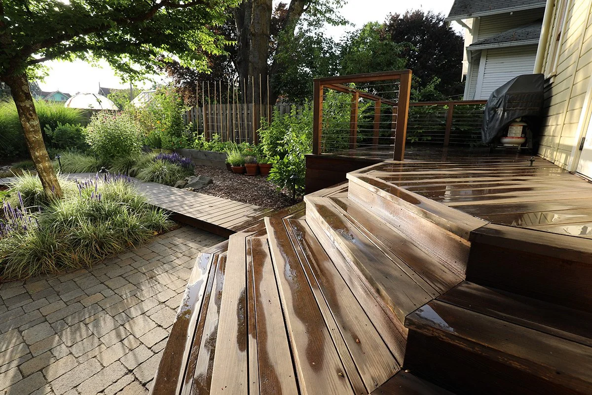 Wet wooden deck stairs leading up to a patio with a grill, overlooking a landscaped garden with trees and plants.
