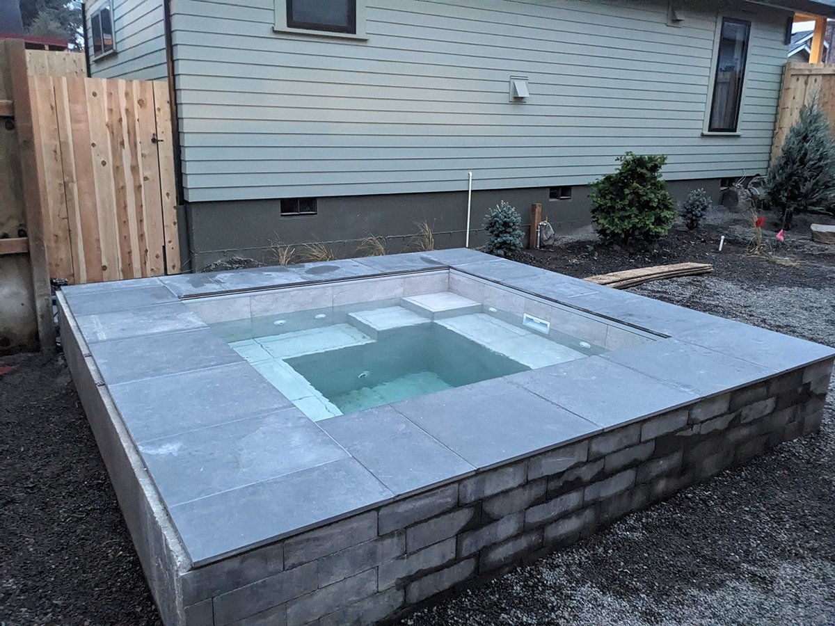 A small rectangular hot tub or spa with stone tiles surrounding it, located in a backyard next to a house with beige siding and a wooden fence.