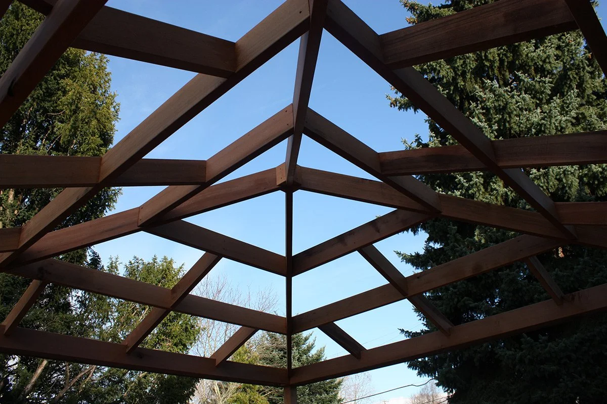 Looking up at a partially constructed wooden gazebo frame with trees and a blue sky in the background.