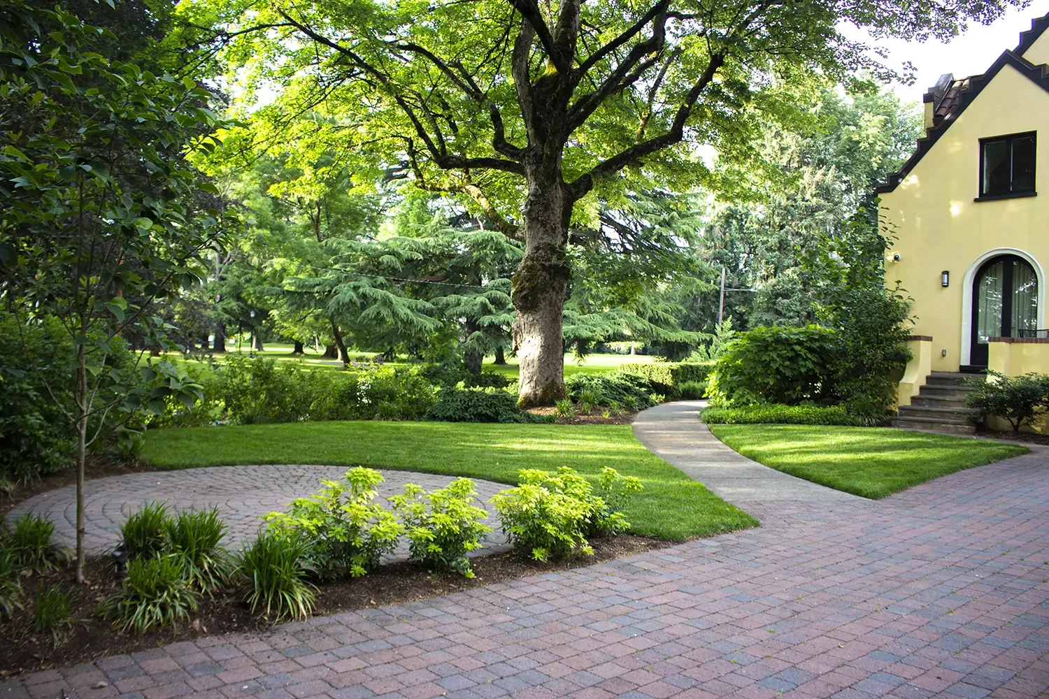A landscaped front yard with a brick pathway, green grass, trees, bushes, and part of a yellow house with steps and a rounded window.