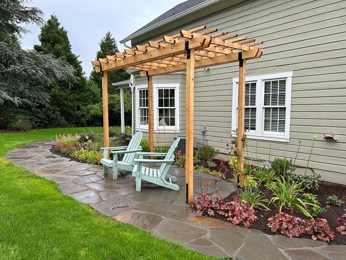 Backyard patio with two light blue Adirondack chairs under a wooden pergola next to a beige house with white trim, surrounded by green grass, plants, and trees.