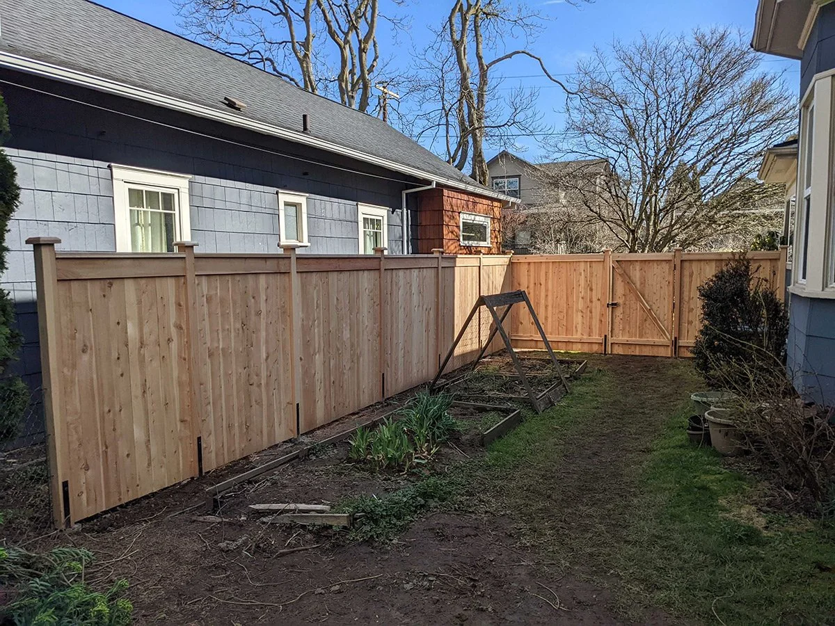 Backyard with a newly installed wooden fence, garden bed, old swing set, and neighboring houses with trees in the background during daytime.