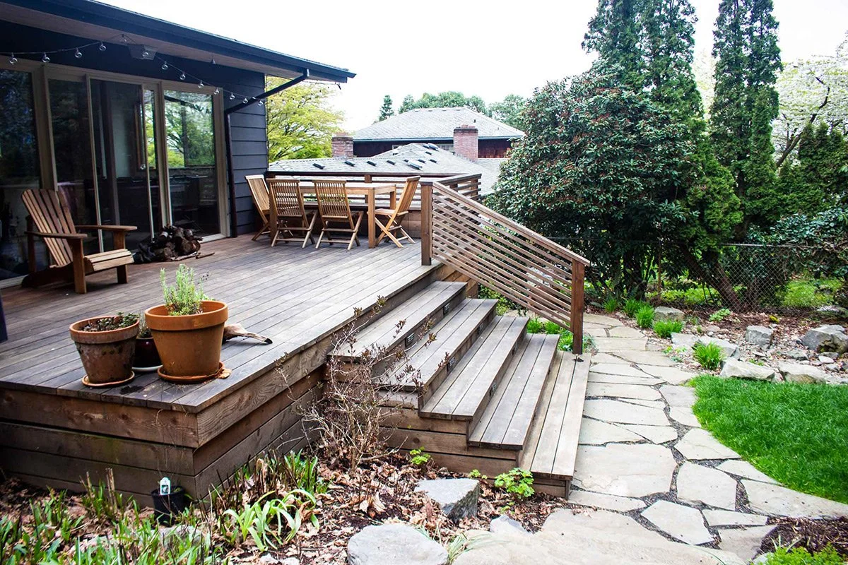 A wooden backyard deck with patio furniture, potted plants, and a garden path surrounded by greenery.