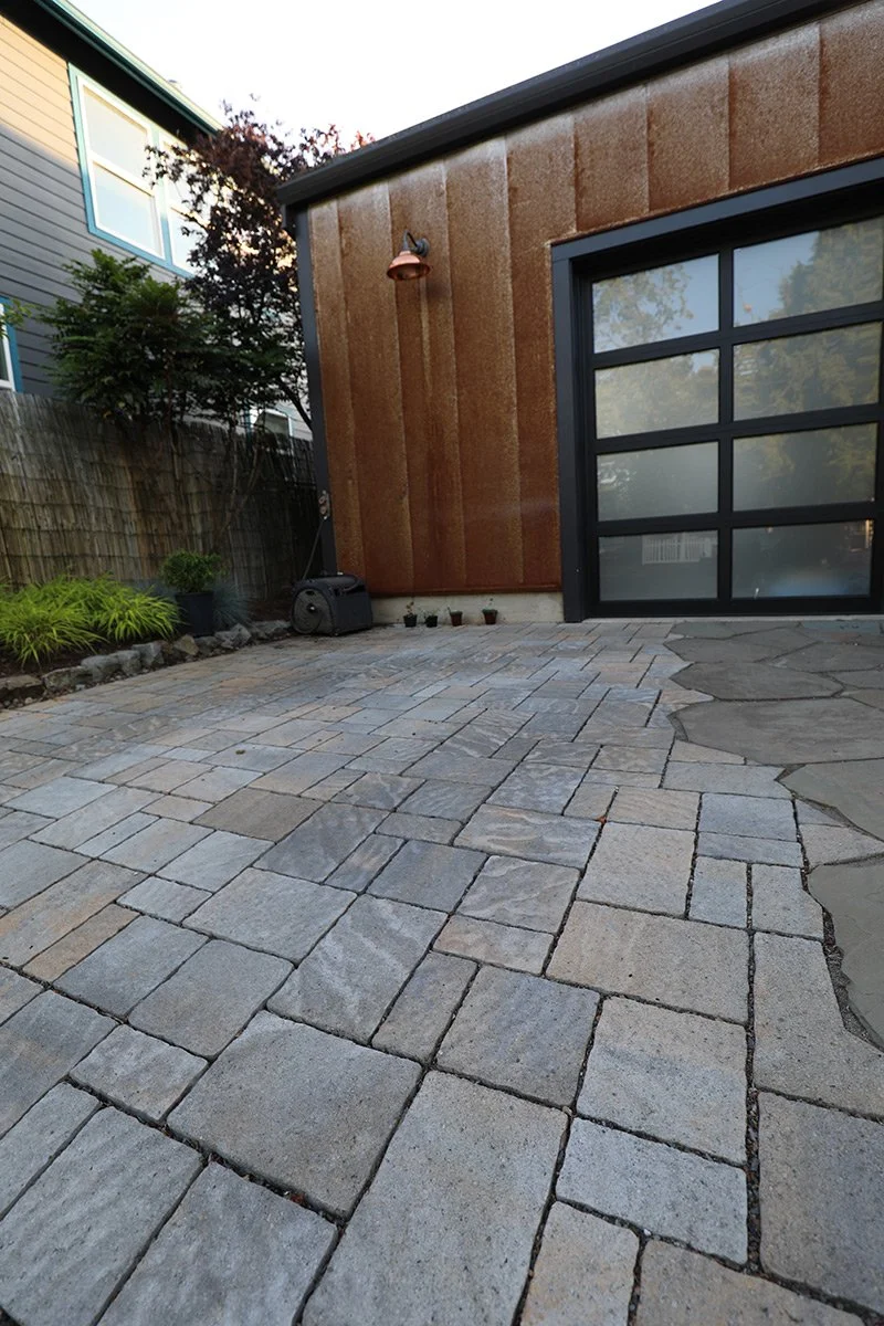 A backyard patio area with a stone tile surface, adjacent to a building with rust-colored siding and large glass garage door. There is a garden bed with plants and a wooden fence nearby, as well as a small black speaker or device on the ground.
