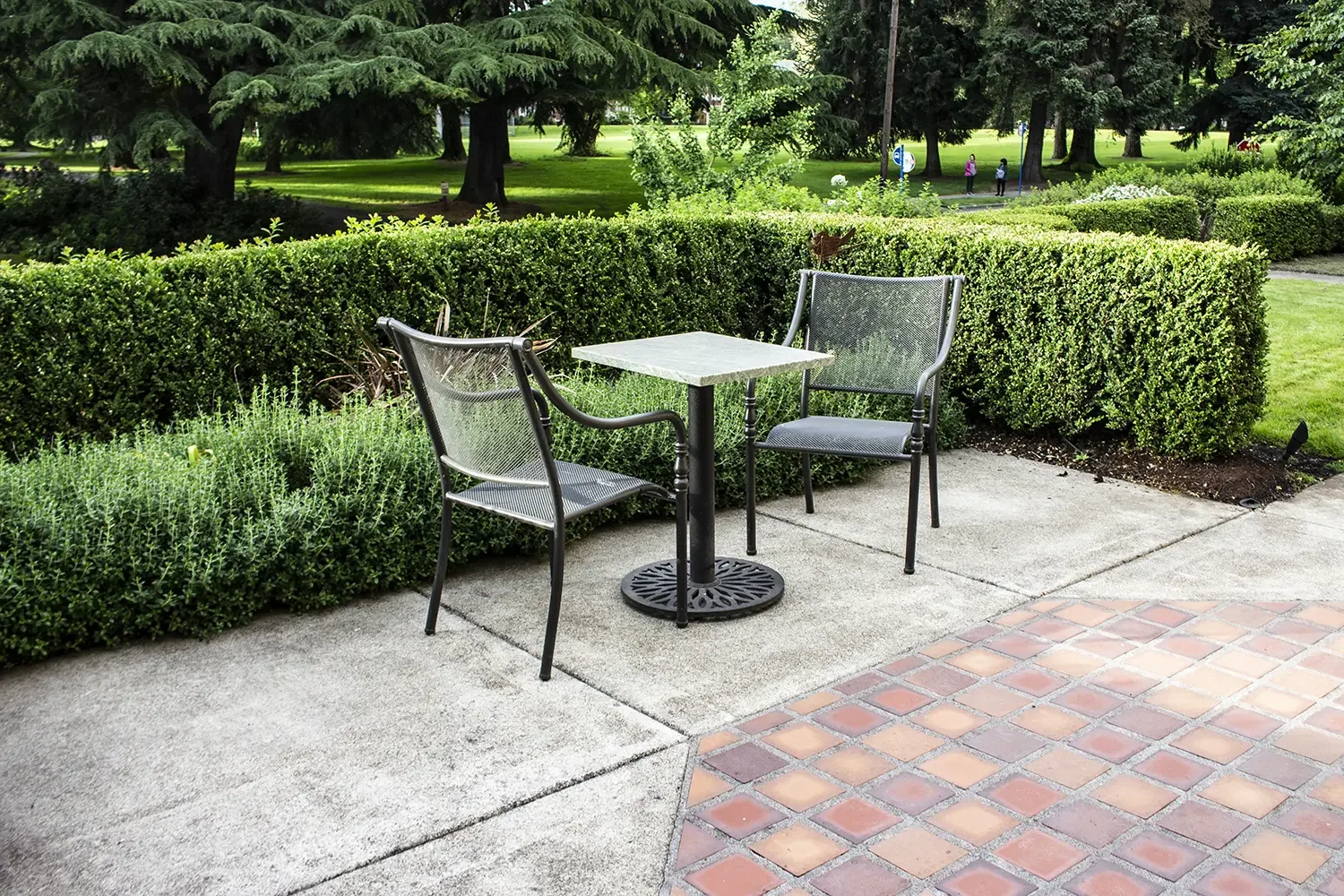 Empty outdoor patio area with two metal chairs and a small square table, surrounded by trimmed bushes and green trees in the background.