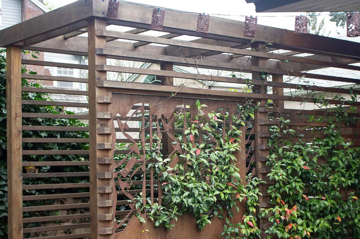 A wooden outdoor pergola with string lights, surrounded by green foliage and vines, with a decorative cut-out metal panel.