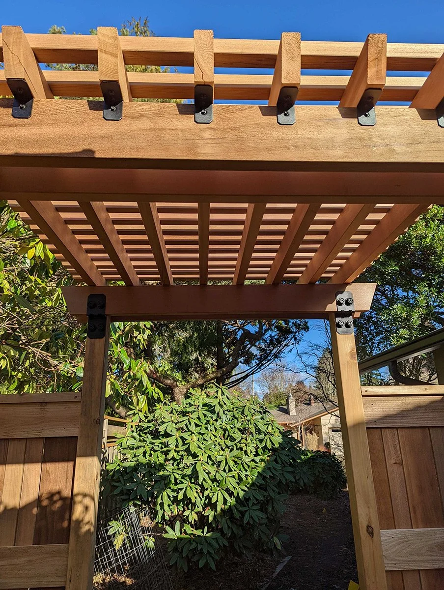 View of a wooden pergola under construction, with a lattice roof and outdoor garden area with greenery and a neighboring house in the background.