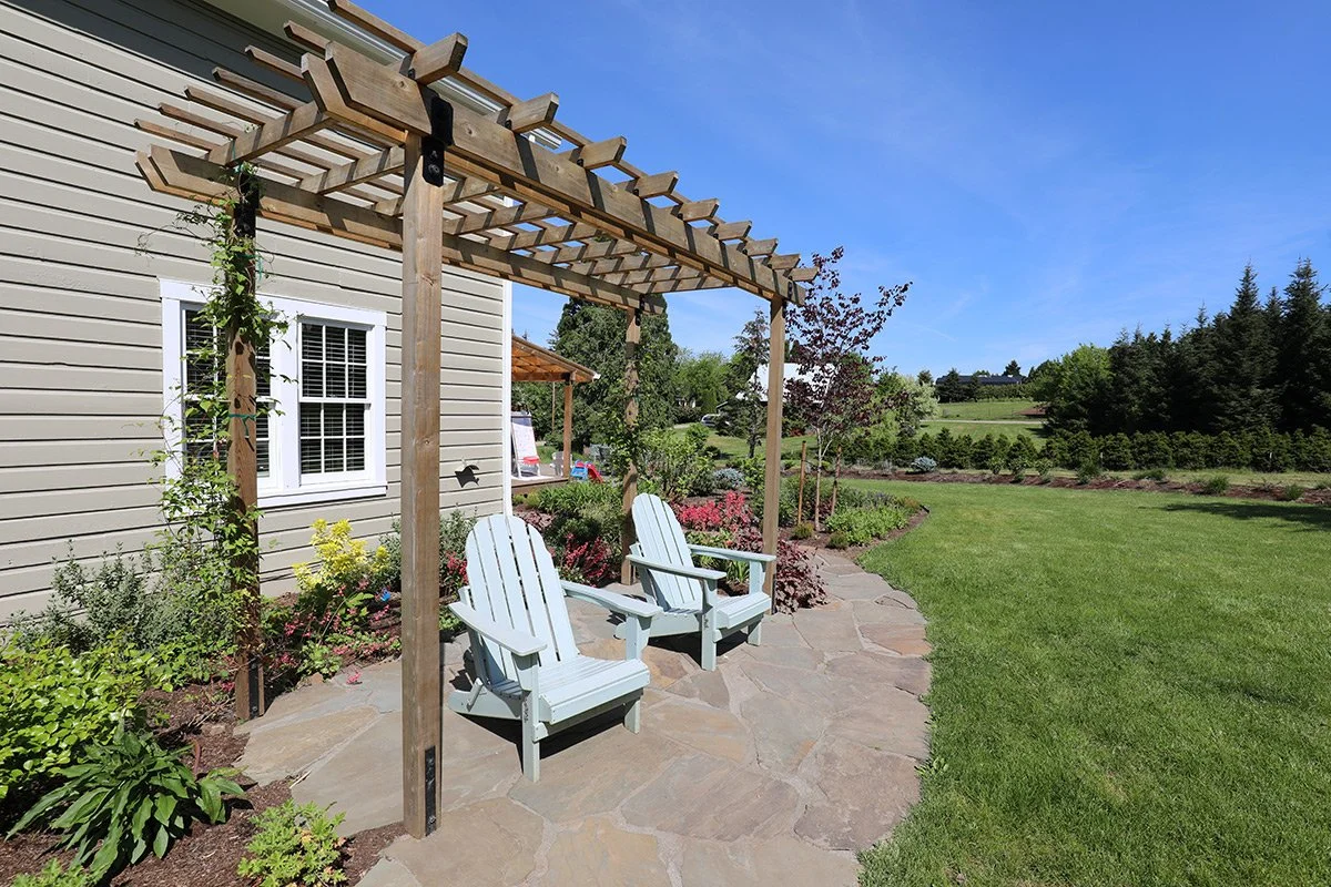 A backyard patio with two light blue Adirondack chairs under a wooden pergola, surrounded by a garden with colorful flowers and lush green lawn, with trees and fields in the background under a blue sky.