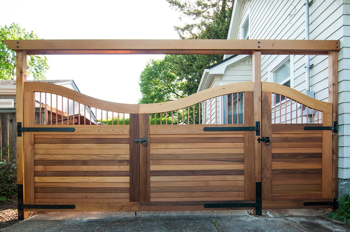 Wooden gate with decorative curved top and black hardware in front of a house with white siding and greenery.