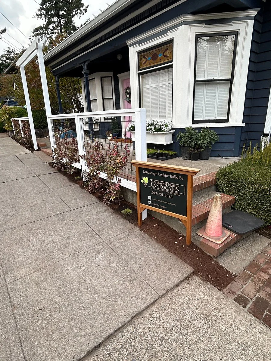 Front porch of a blue house with white trim, decorated with potted plants and a flower box, and a signboard advertising landscape design services.