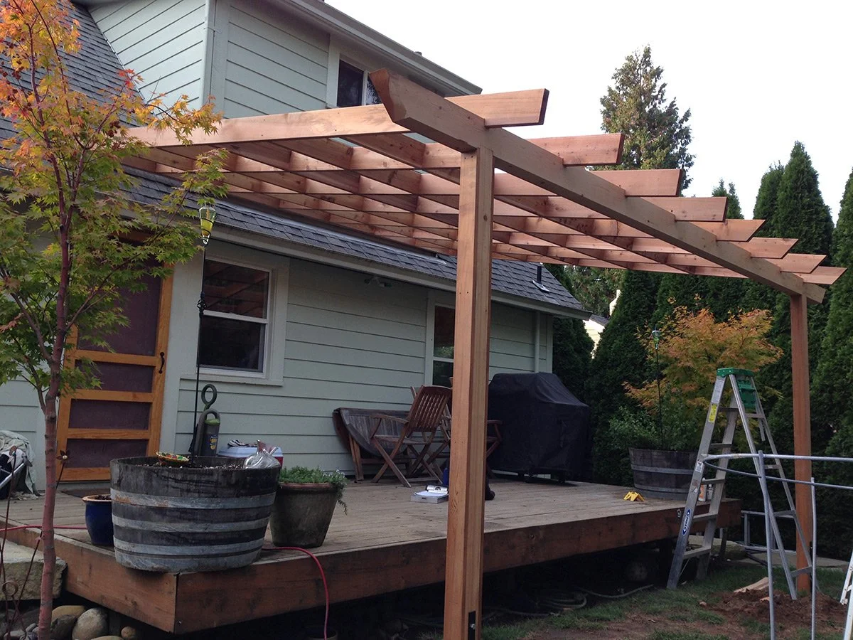 Backyard deck with wooden pergola under construction, ladder, chairs, potted plants, and grill.