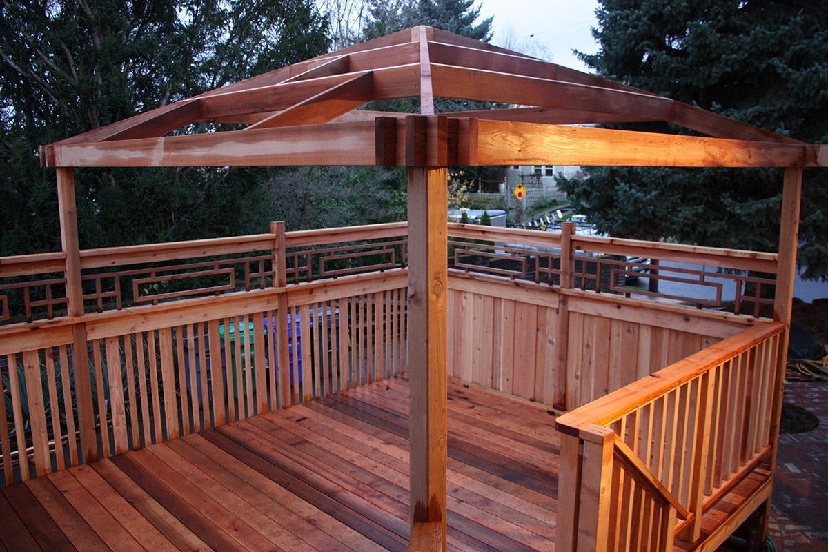 A wooden deck with an open framework gazebo structure, surrounded by a wooden railing, with trees and a street with parked cars in the background.