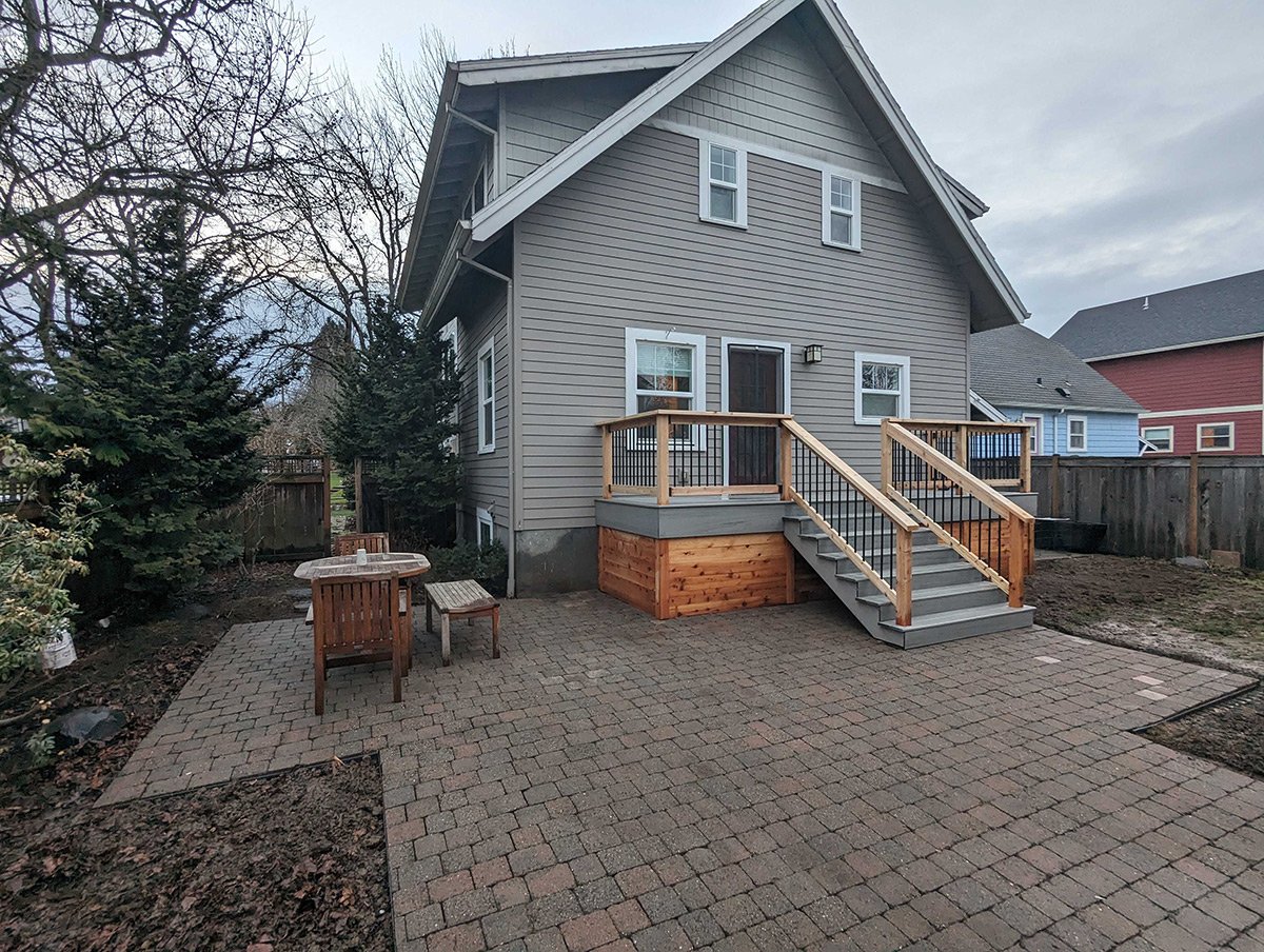 Backyard with a newly built wooden deck with stairs, leading to a gray house with beige siding, surrounded by a brick patio and some trees.