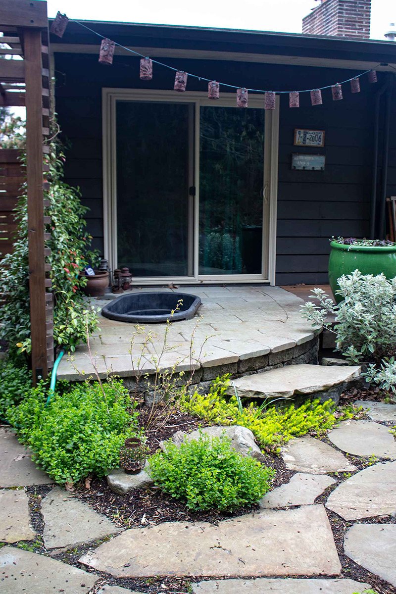 Backyard patio with a stone stepping stone pathway, small bushes, a round hot tub, and a sliding glass door leading into a house, decorated with a string of hanging lanterns.
