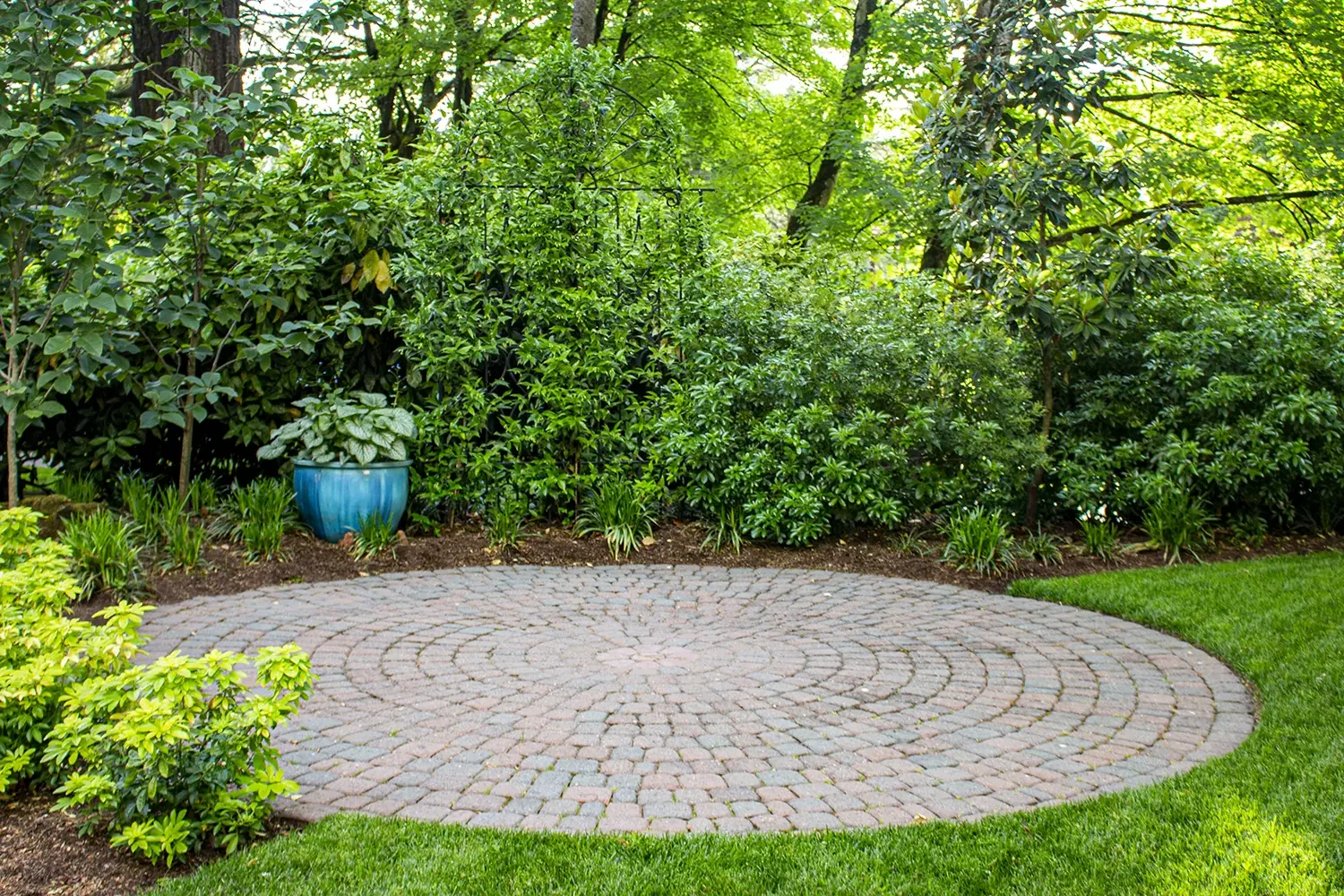 A circular brick patio surrounded by lush green bushes and trees, with a blue ceramic pot containing a leafy plant placed on the edge of the patio.