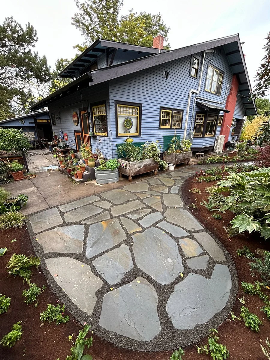 A blue house with a red brick chimney, multiple yellow-framed windows, and a garden with potted plants and a stone pathway surrounded by mulch.