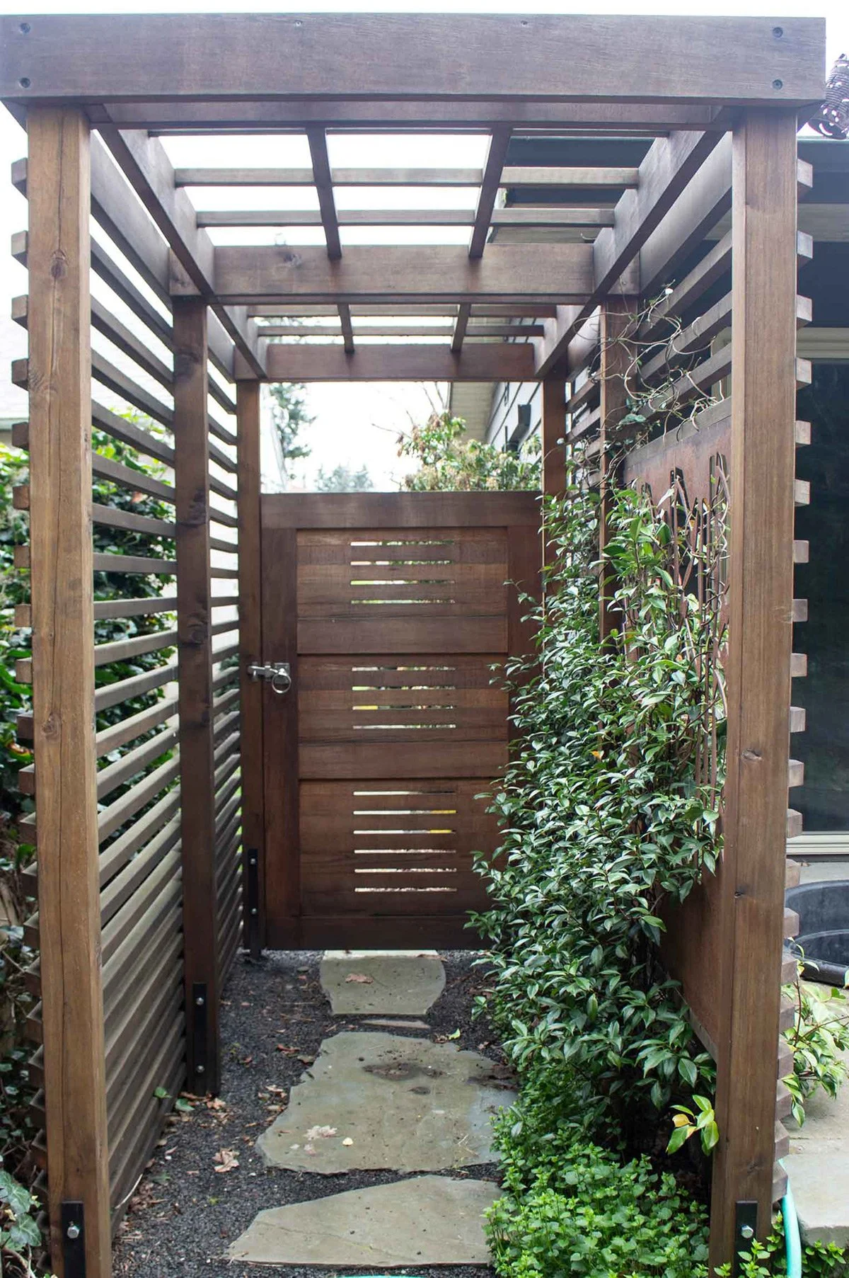 Wooden outdoor privacy structure with a small gate, pathway, and climbing plants.