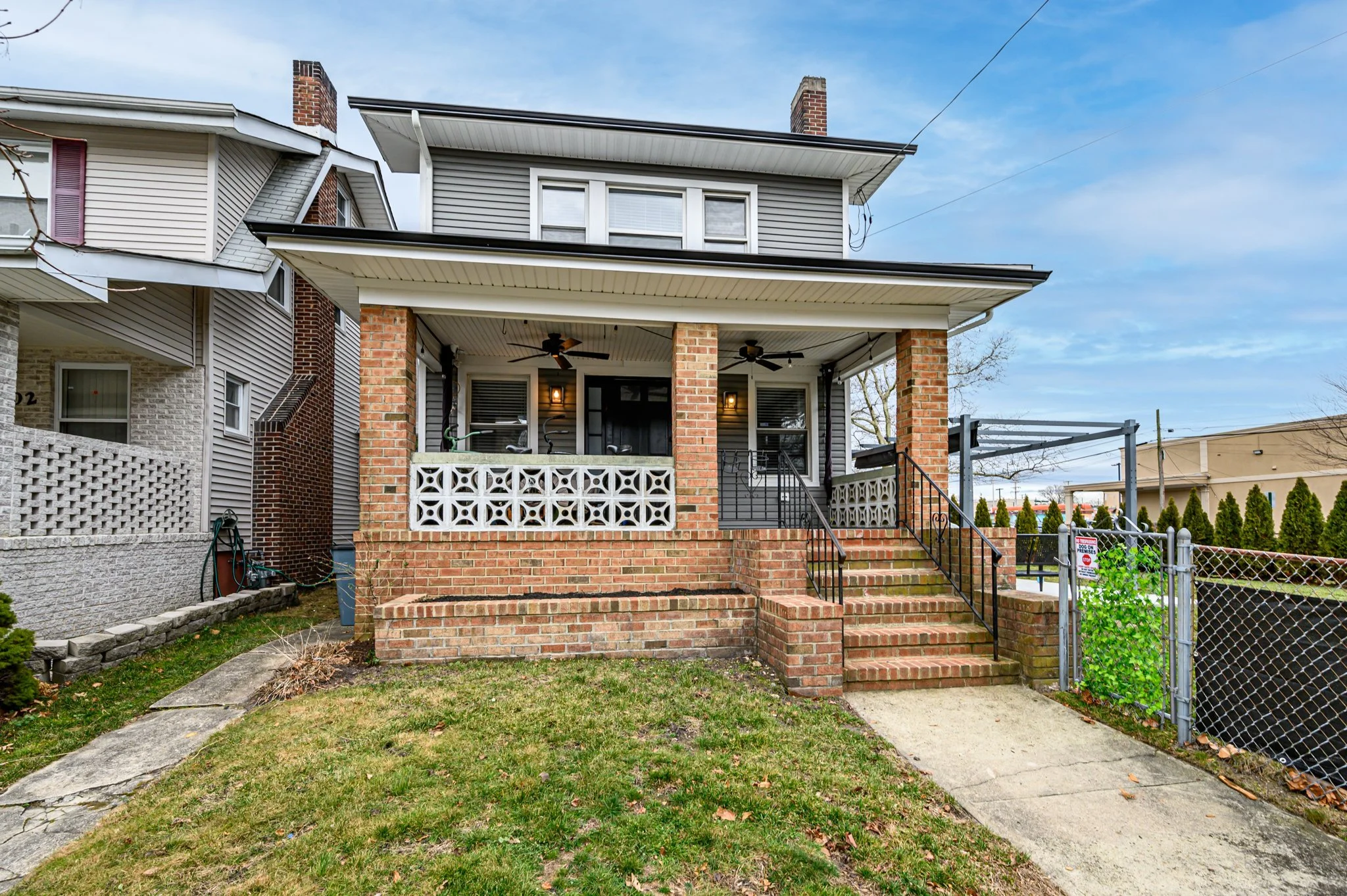 Front view of a brick house with a porch, stairs, and black railings, with neighboring houses, a grassy lawn, and a chain-link fence.