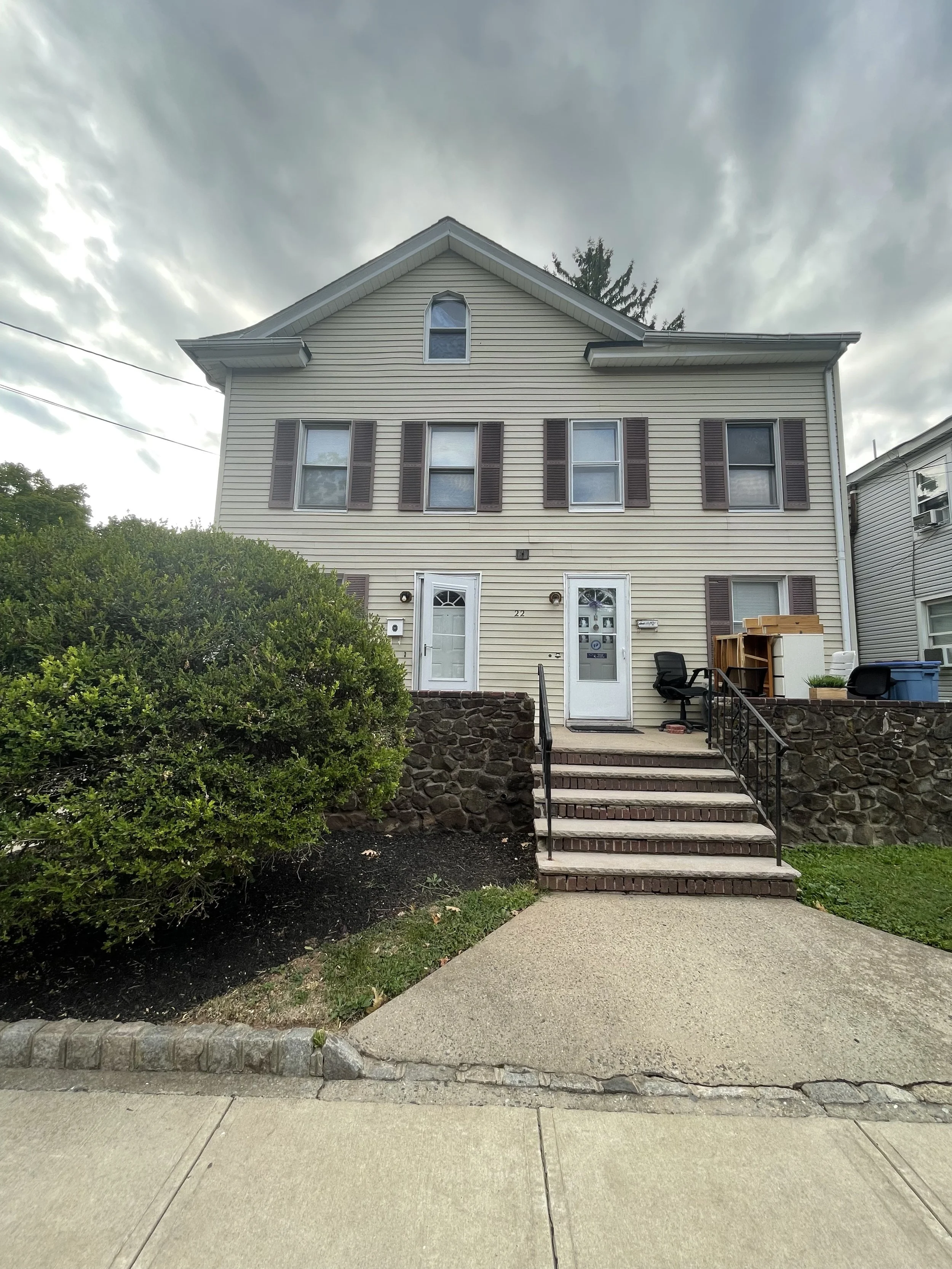 Front view of a beige three-story house with brown shutters, steps leading to two doors, and a cloudy sky overhead.