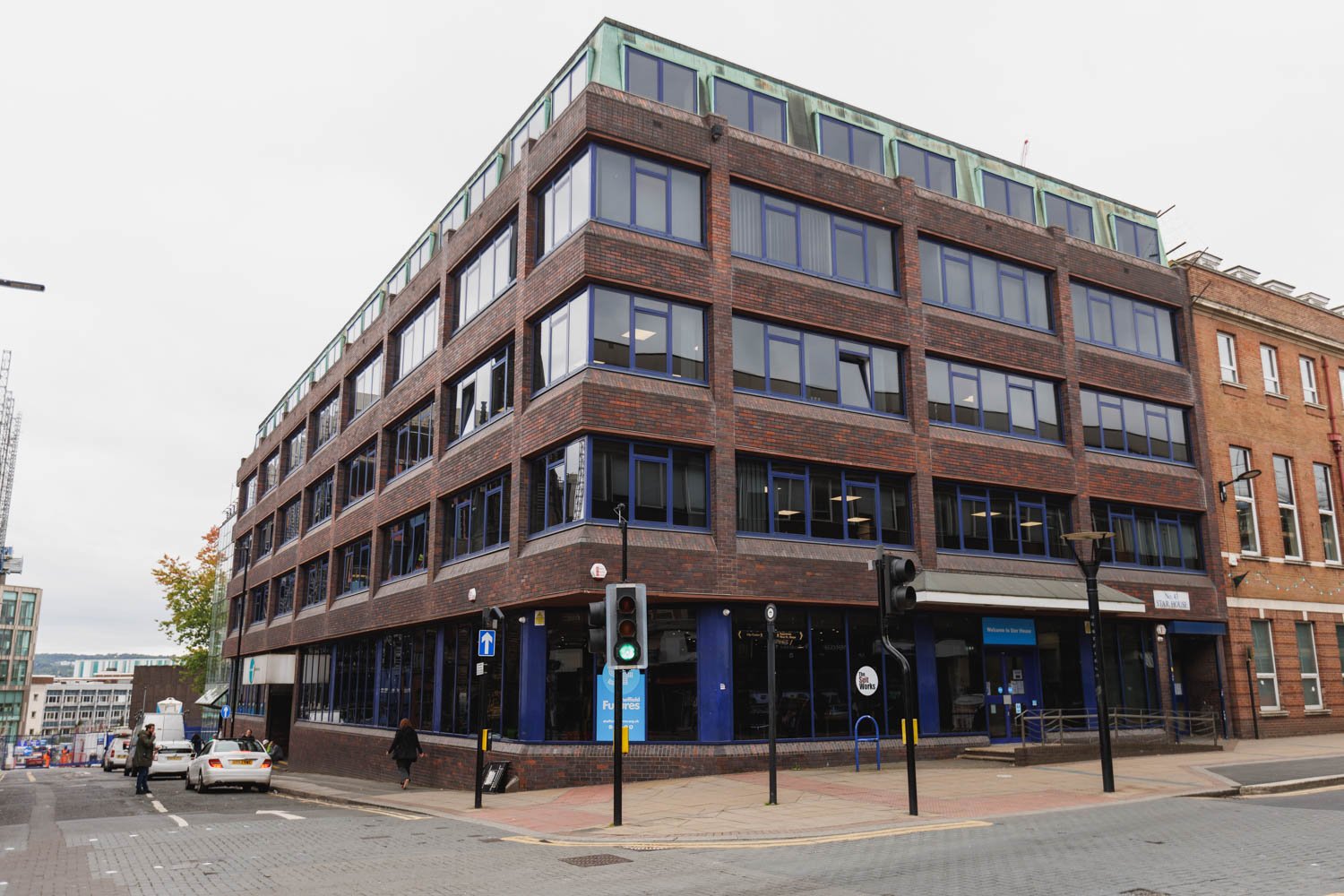 Multi-story brick office building with large windows on a street corner, traffic lights, pedestrians, and parked cars.