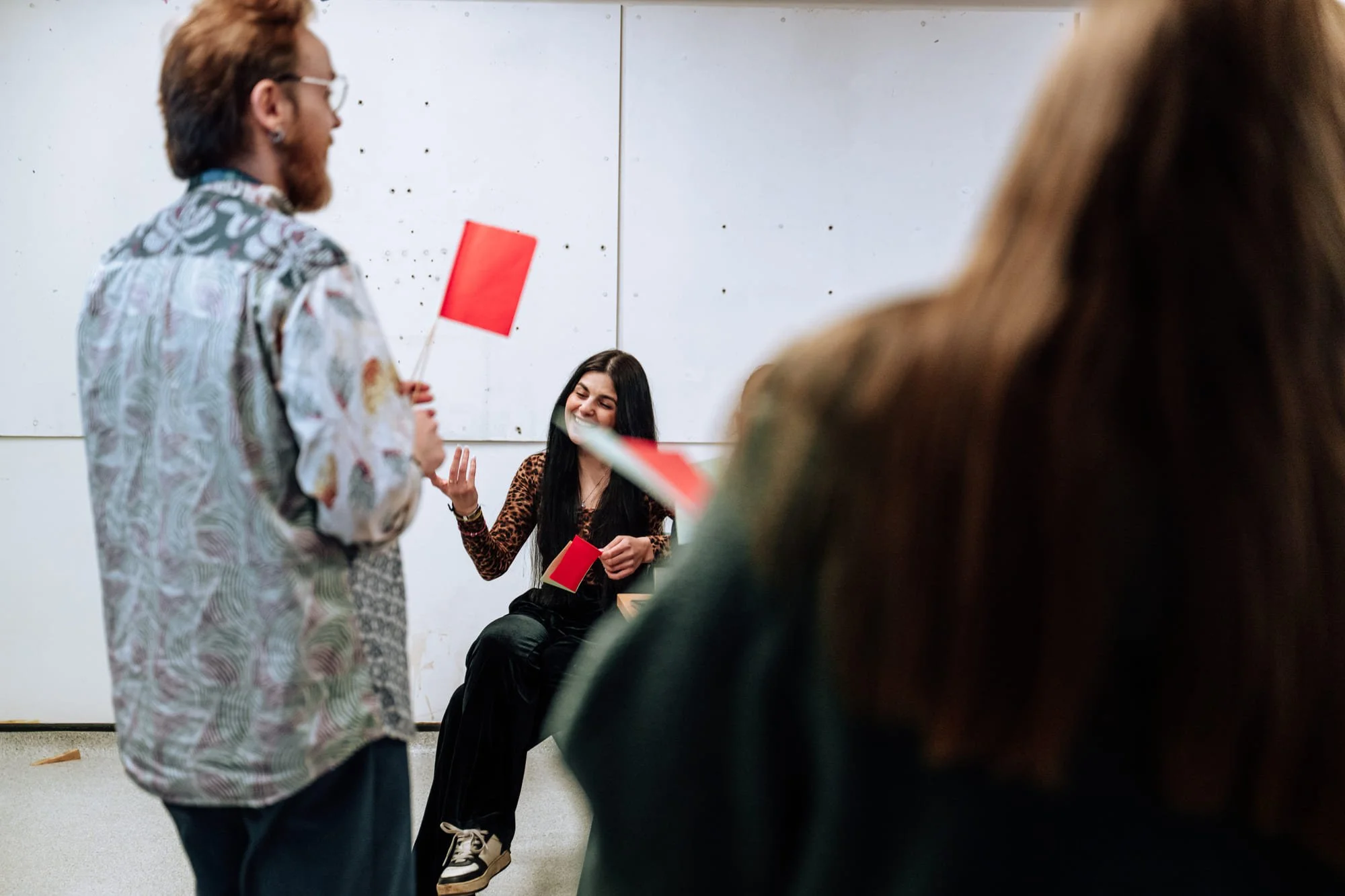 A woman with long black hair sitting and smiling during a group activity, with two people partially visible in the foreground, one holding a red flag, in a white room.