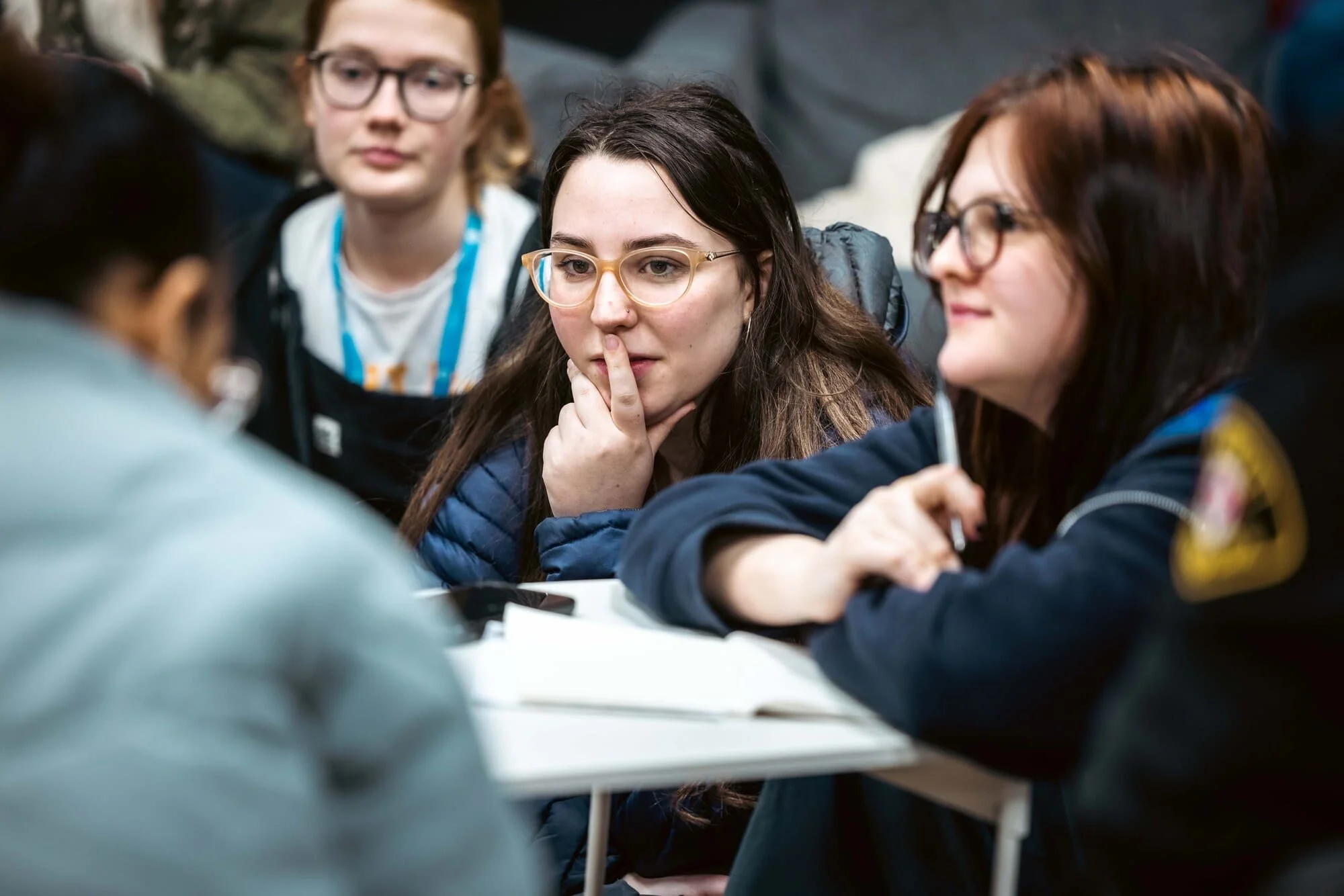 A group of young women sitting at a table, engaged in conversation or a discussion, some taking notes. The women are wearing glasses and casual clothing.