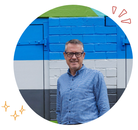 A man in glasses and a blue checkered shirt smiling in front of a colorful painted brick wall with blue, white, black, and green sections.