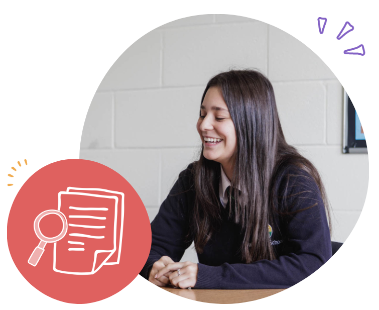 A young woman with long dark hair, smiling and sitting at a table in a classroom or office, in front of a light gray brick wall.