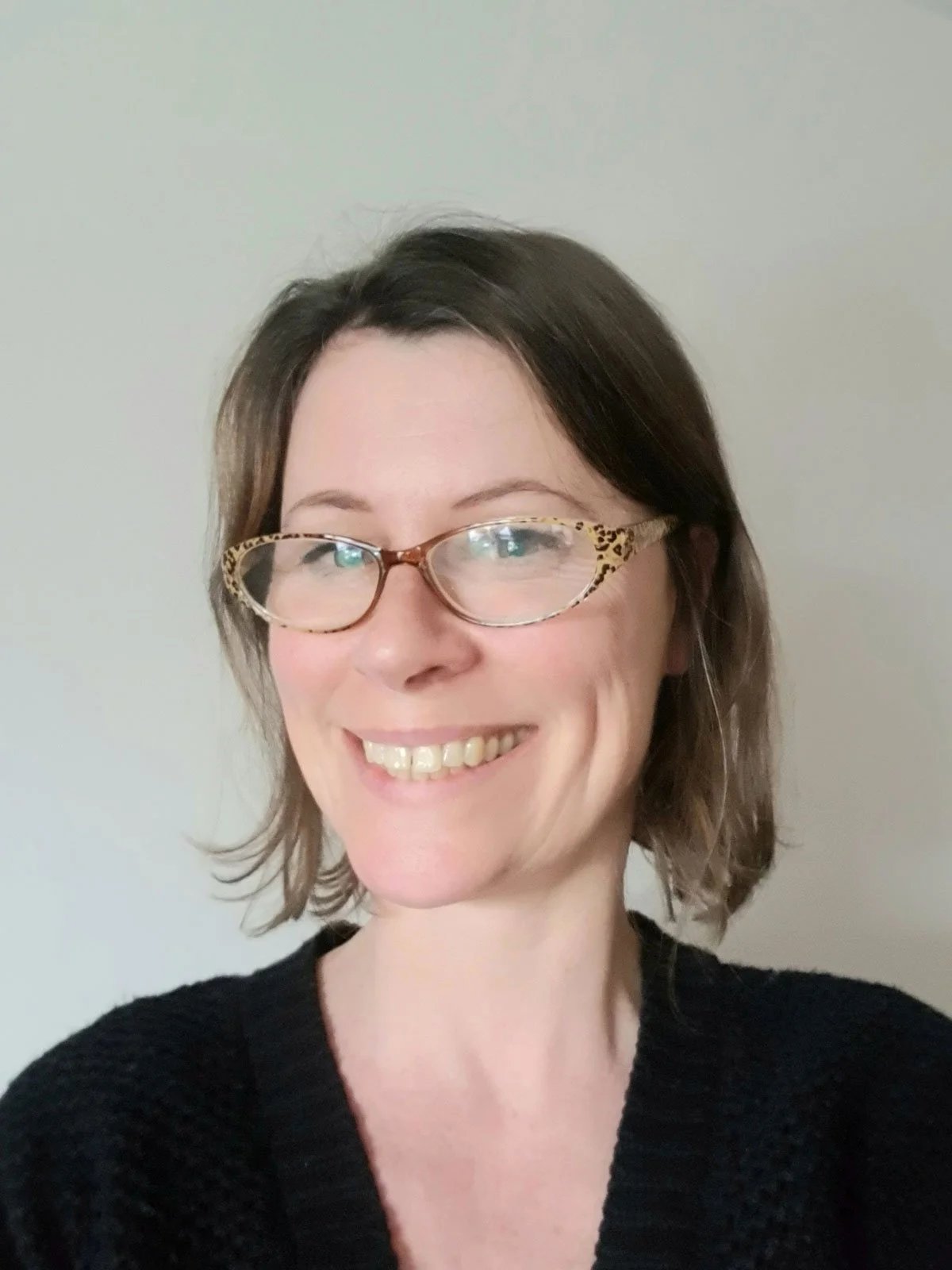 Smiling woman with short brown hair and glasses with a leopard print pattern, wearing a black sweater, standing against a plain light-colored background.