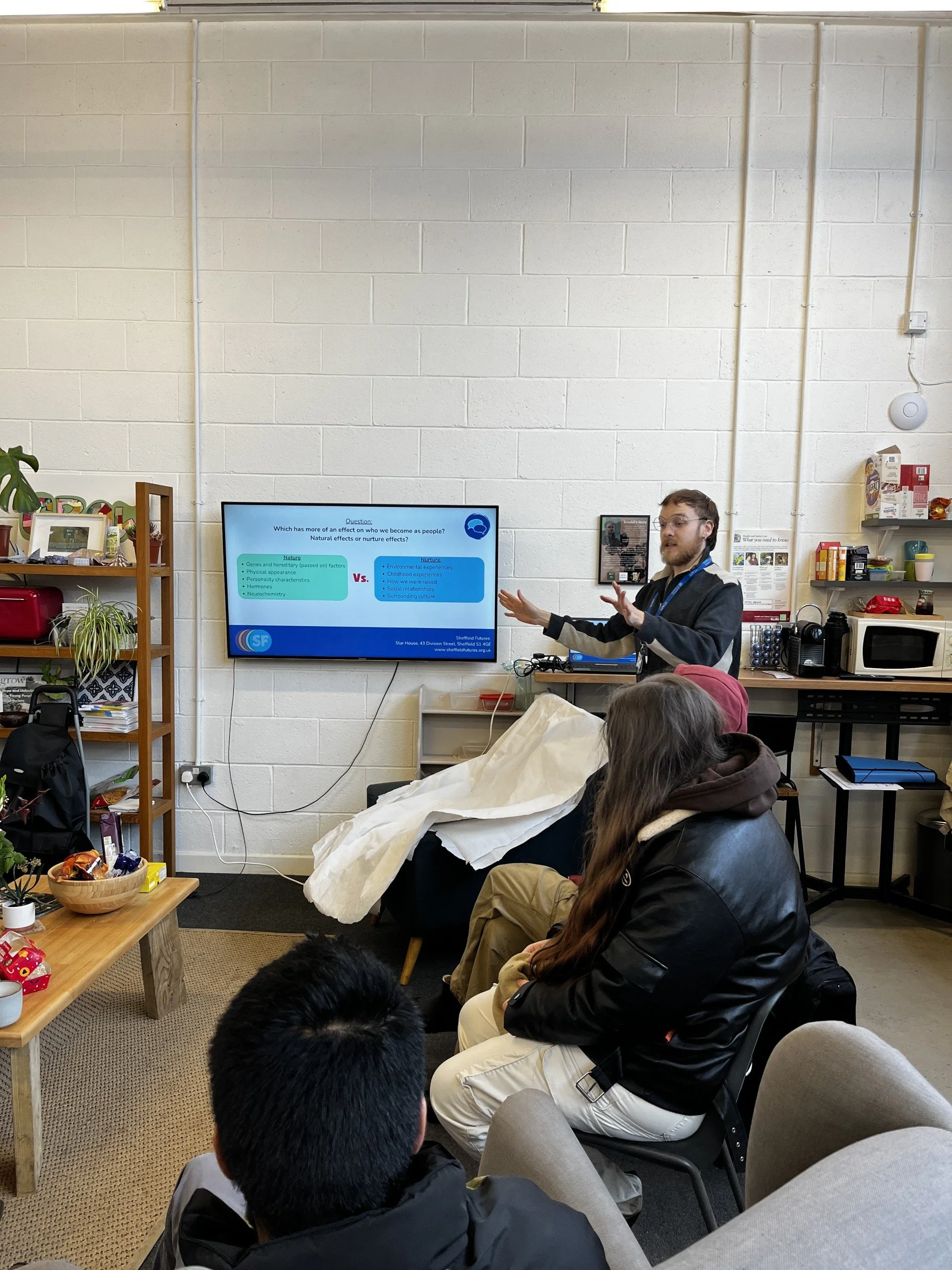 A man giving a presentation to a small group in a casual office or classroom setting. The presenter is near a large screen displaying a slide, and several people are seated, listening attentively.