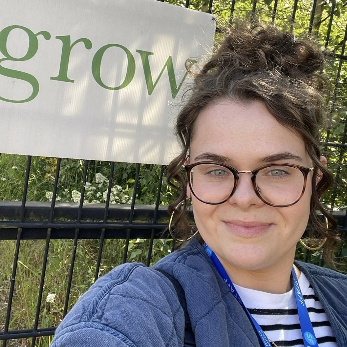 Self-portrait of a woman with glasses and curly hair, smiling outdoors in front of a black fence with plants and a sign that partially reads 'grow'.