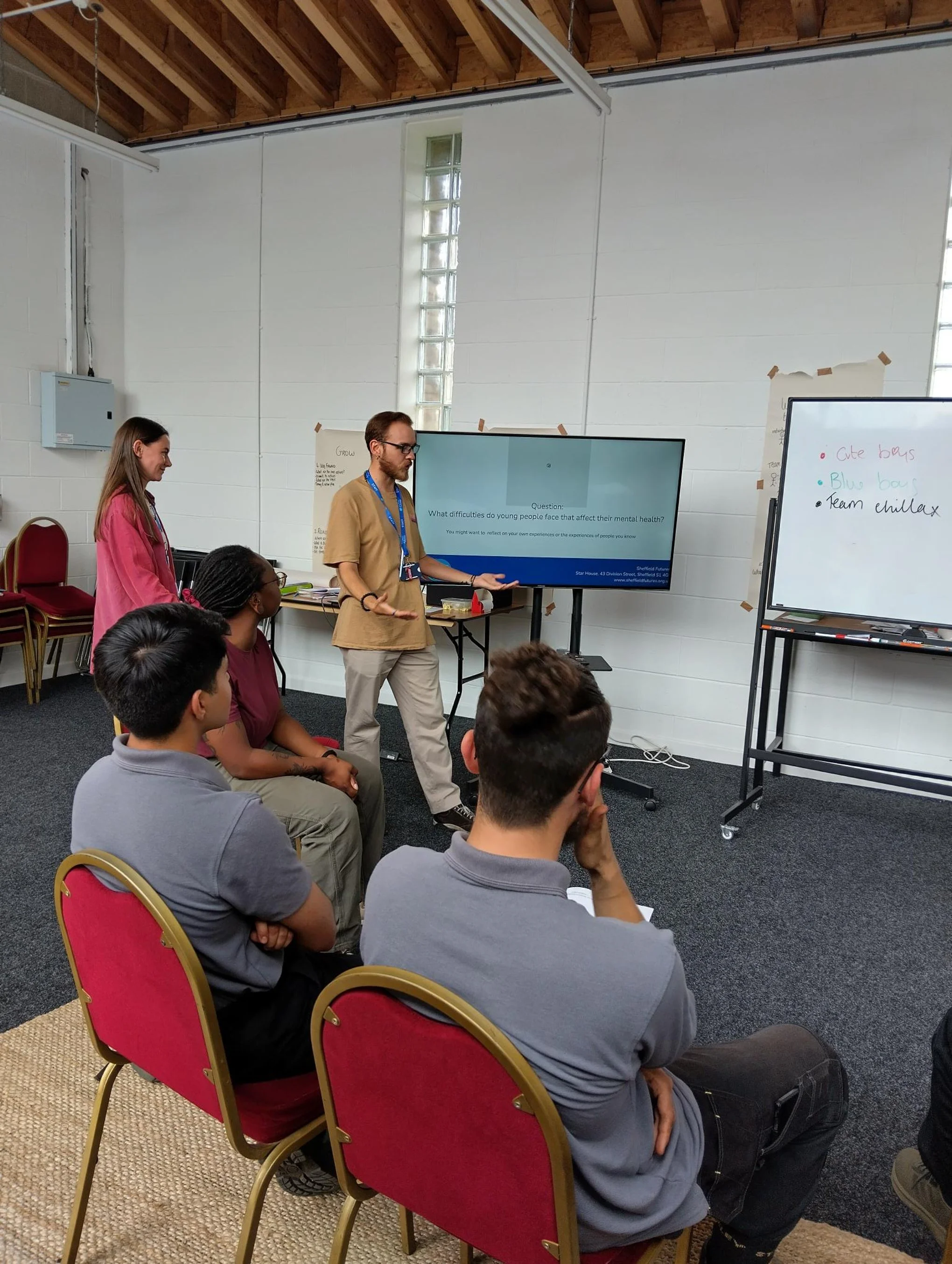 A group of people attending a presentation in a room with white walls. Two presenters stand near large screens displaying slides, while five audience members sit facing them. The room has a high ceiling with visible wooden beams and a small window with glass blocks.