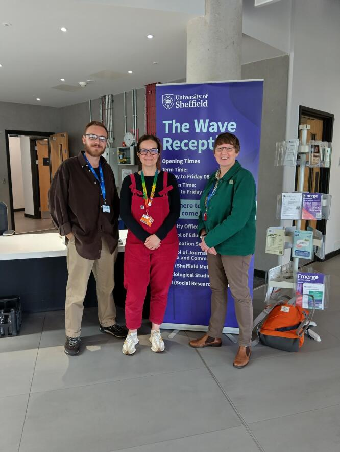 Three people standing indoors in front of a sign that reads 'The Wave Reception' at the University of Sheffield, with a reception desk and literature stands nearby.