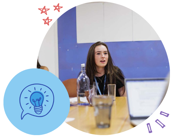 Young woman with long dark hair sitting at a table during a meeting or discussion, with glasses and a water bottle in front of her, in a room with blue and white walls.